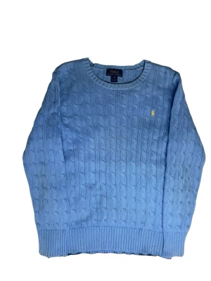 Ljusblå kabelstickad tröja Polo Ralph Lauren - Ljusblå kabelstickad tröja från Polo Ralph Lauren med rund halsringning och klassisk gul broderad logga på bröstet. Tröjan har ribbade muddar och är tillverkad i mjukt material som ger en skön passform.