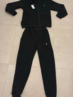 Svarta mjukisbyxor från Polo Ralph Lauren - Svarta mjukisbyxor från Polo Ralph Lauren med klassisk logga broderad i vitt på benet. Byxorna har snörning i midjan och är gjorda i mjukt bomullsmaterial med ribbade muddar vid bensluten. Perfekta för chill och sportiga dagar.