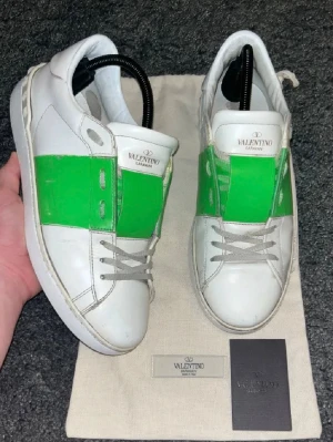 Valentino vita sneakers med grön detalj - Säljer ett par vita sneakers från Valentino Garavani med en bred grön rand över sidan. Skorna har klassisk låg profil, vita snören och är tillverkade i slätt läder. Perfekta för dig som vill sticka ut med en färgklick i din outfit.
