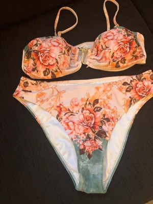 Blommig bikini med rosa rosor - Snygg bikini med blommigt mönster i rosa och gröna toner. Överdelen har justerbara axelband samt guldfärgad detalj framtill. Underdelen har hög midja och samma romantiska blommönster. Perfekt för strandhäng eller poolparty!