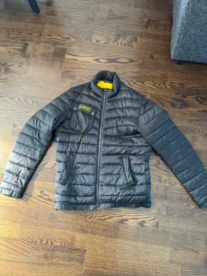 Svart pufferjacka från Barbour - Säljer en svart pufferjacka från Barbour med gul insida och Barbour-logga på bröstet. Jackan har dragkedja framtill, två sidofickor och en innerficka med dragkedja. Perfekt för kalla dagar och har en modern, quiltad design.