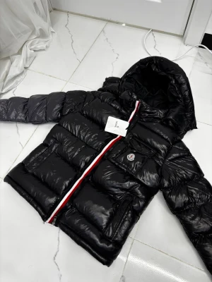 Moncler Maya - Säljer en Moncler Maya dunjacka i svart, storlek S/M. En av Monclers mest ikoniska och eftertraktade modeller med blank finish och huva. Jackan har klassiska Moncler-detaljer såsom logga på bröstet samt dragkedja med röd/vit/blå rand.  Jackan är i mycket fint skick och originaltagg sitter kvar. Varm men samtidigt lätt, perfekt för både vardag och mer uppklädda tillfällen under höst och vinter.  Endast seriösa köpare. Hör av er vid intresse.