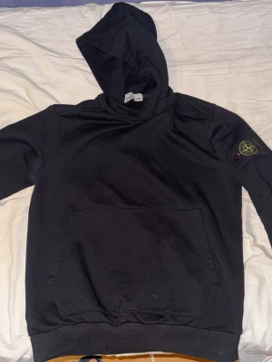 Svart hoodie från Stone Island - Säljer en svart hoodie från Stone Island med klassisk patch på vänster ärm. Hoodien har stor ficka framtill och ribbade muddar. Tillverkad i mjuk bomull för skön känsla och avslappnad stil. Perfekt för dig som gillar streetwear och vill ha något stilrent.