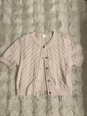 Ljusrosa kabelstickad kofta Gina Tricot - Söt ljusrosa kabelstickad kofta från Gina Tricot med korta ärmar och v-ringning. Koftan har ribbade muddar och knäpps med fyra knappar framtill. Perfekt att slänga över en topp för en trendig look.