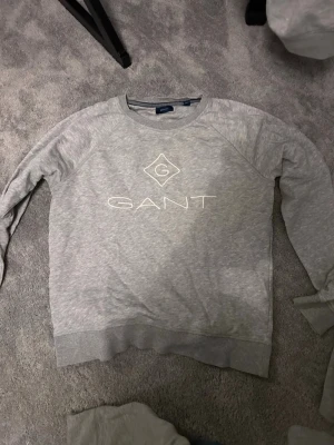 Grå sweatshirt från GANT - Snygg grå sweatshirt från GANT med vit logga och emblem på bröstet. Tröjan har rund halsringning, långa ärmar och ribbade muddar vid ärmslut och nederkant. Perfekt för en avslappnad och stilren look.