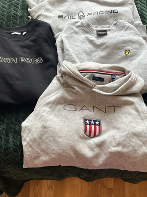 Sweatshirtpaket GANT, Sail Racing, Björn Borg, Lyle & Scott  - Fyra snygga sweatshirts i bomull från GANT, Sail Racing, Björn Borg och Lyle & Scott. Tre grå och en svart, alla med broderade eller tryckta loggor framtill. Perfekta för en avslappnad och sportig stil. GANT tröjan har ett litet hål, se bild.