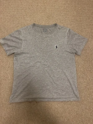 Grå t-shirt från Ralph Lauren - En grå t-shirt från Ralph Lauren med klassisk passform och liten broderad logga på bröstet. T-shirten har rund halsringning och korta ärmar, perfekt för en avslappnad look. 