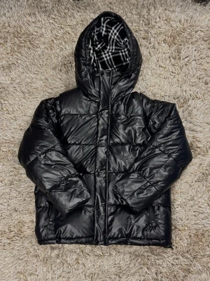 Svart pufferjacka från Burberry - Snygg svart pufferjacka från Burberry med klassiskt rutigt foder i huvan. Jackan har dragkedja, två fickor och en vadderad design som håller dig varm. Perfekt för kalla dagar och har en modern, boxig passform.