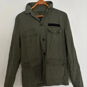 Olivgrön overshirt med fickor - Snygg olivgrön overshirt med fyra stora fickor framtill och klassisk krage. Jackan har knäppning med knappar och en svart detalj på bröstfickan. Perfekt för lager på lager och en avslappnad stil. Längd: 70cm, P2P: 49cm