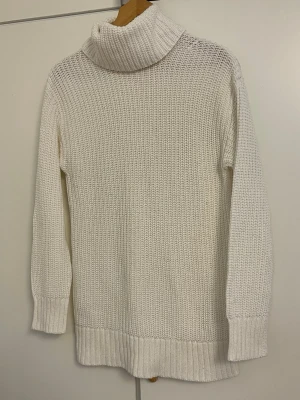 Vit polotröja i grovstickat - Snygg vit stickad polotröja med grov struktur och långa ärmar. Tröjan har en hög, bred polokrage och ribbstickade muddar vid ärmslut och nederkant. Perfekt för kalla dagar och enkel att matcha med jeans eller kjol.