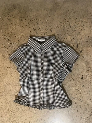 Rutig blus från Princess Polly - Supersöt svartvit rutig blus från Princess Polly med korta ärmar och knappar framtill. Blusen har en markerad midja, volangkant nertill och klassisk krage. 
