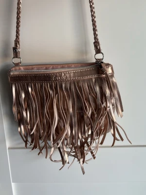 Kuvertväska i metallic med fransar - Säljer en snygg kuvertväska från Lively i metallic roséguld. Väskan har långa fransar framtill och en flätad axelrem. Tillverkad i glansigt konstläder och stängs med dragkedja. Perfekt för dig som vill sticka ut med en trendig accessoar.