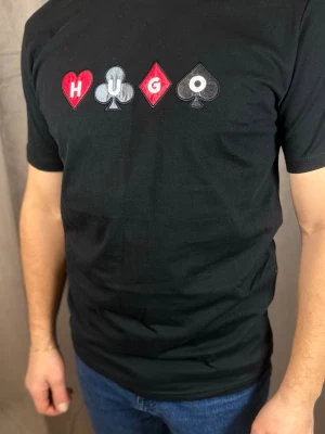 Svart HUGO t-shirt med spelkortstryck - Svart t-shirt från HUGO med coolt tryck av spelkortssymboler och bokstäver på bröstet. T-shirten har rund hals och korta ärmar. Materialet känns mjukt och skönt, troligtvis bomull. Perfekt för dig som gillar unika prints och streetstyle.
