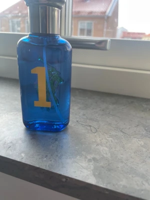 Ralph Lauren Polo Blue parfym - Fräsch parfym från Ralph Lauren i en blå glasflaska med silverfärgat lock. Flaskan har en stor gul etta på framsidan och en grön polospelare i bakgrunden. Perfekt för dig som gillar sportiga och fräscha dofter.