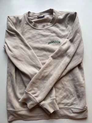 Beige sweatshirt från Clique - Beige sweatshirt med rund hals och tryck i grönt på bröstet och ryggen där det står 'Jameson Irish Whiskey'. Tröjan har långa ärmar och ribbade muddar vid ärmslut och nederkant. Perfekt för dig som gillar stilrena och avslappnade plagg.