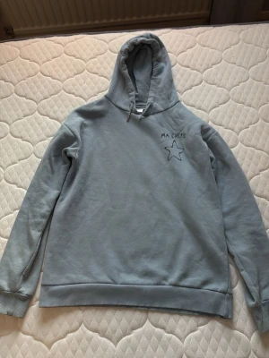 Ljusblå hoodie från Gina Tricot - Ljusblå hoodie från Gina Tricot med broderad text 'MA CHERIE' och en stjärna på bröstet. Klassisk modell med huva och dragsko samt ribbade muddar vid ärmslut och nederkant. Perfekt för en chill och avslappnad stil.