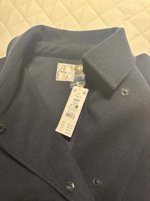 Mörkblå cape- poncho-kappa från Gina Tricot - Stilren mörkblå kappa från Gina Tricot med klassisk krage och knäppning framtill. Kappan har en enkel och tidlös design som passar perfekt till höst och vinter. Materialet känns mjukt och har en matt finish. Snygg att matcha med både jeans och kjol.