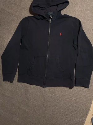 Svart hoodie från Polo Ralph Lauren - Klassisk svart hoodie från Polo Ralph Lauren med dragkedja framtill och två fickor. Ikoniska röda loggan broderad på bröstet. Tillverkad i mjukt bomullsmaterial och har en skön huva. Perfekt för en avslappnad och stilren look.