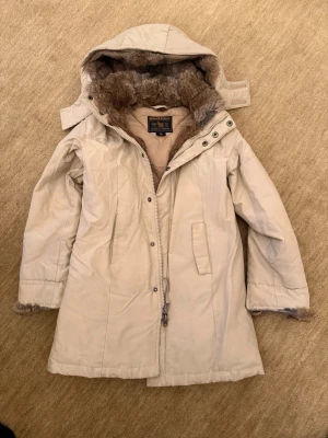 Beige parkas med fuskpäls från Woolrich - Snygg beige parkas från Woolrich med avtagbar huva och mjukt fuskpälsfoder. Jackan har dragkedja och knappar framtill, två fickor och pälsdetaljer vid ärmslut och huva. Perfekt för kalla dagar och sopren design.