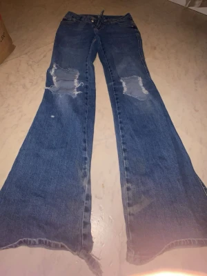 Blå utsvängda jeans med slitningar - Säljer ett par blå jeans med utsvängda ben och slitna detaljer låren. Jeansen har råa kanter nertill och klassisk femficksmodell. Perfekta för dig som gillar en avslappnad och trendig stil.