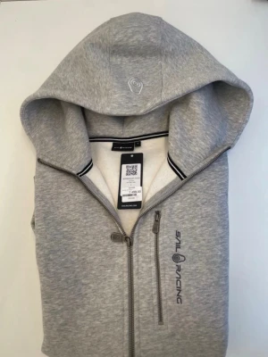 Grå hoodie från Sail Racing - Snygg grå hoodie från Sail Racing med dragkedja hela vägen, huva och logga på bröstet. Hoodien har en bröstficka med dragkedja och ribbade muddar vid ärmslut och nederkant. Perfekt för en sportig och avslappnad stil.