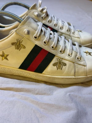 Gucci Ace sneakers – Svart – Guldstjärnor & bin – Strl 41 – Sjukt bra skick - Sjukt snygga Gucci Ace sneakers med guldiga stjärnor och bin, storlek 41, i sjukt bra skick. En riktig statement-sko som sticker ut direkt men fortfarande är stilren.  Perfekta för dig som gillar high-end streetwear, Y2K eller fashion-forward outfits.  • Märke: Gucci • Modell: Ace • Detaljer: Guldstjärnor & bin • Storlek: 41