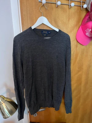 Mörkgrå stickad tröja Polo Ralph Lauren - Mörkgrå stickad tröja från Polo Ralph Lauren med rund halsringning och diskret broderad logga på bröstet. Tröjan har långa ärmar och ribbade muddar vid ärmslut och nederkant. Perfekt för lager på lager och stilren look.