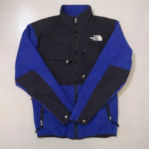 Blå fleecejacka från The North Face - Blå och svart Denali fleecejacka från The North Face med hög krage, dragkedja framtill och flera praktiska fickor. Jackan har logga på bröstet och ryggen samt justerbara muddar. Perfekt för kyliga dagar och outdoor-äventyr.