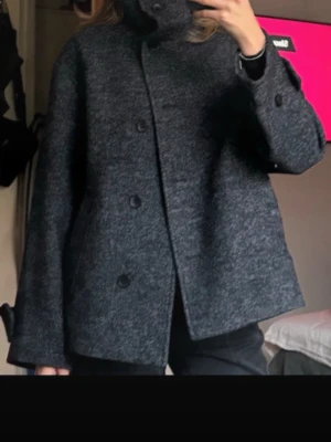 Mörkgrå ullrock med hög krage - Säljer en mörkgrå rock/kappa  i ullmix med hög krage och stora knappar framtill. Kappan har en boxig, rak passform och två sidofickor.