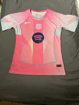 Nike Barcelona rosa matchtröja - Rosa Barcelona matchtröja från Nike med klubbmärke och sponsortryck på bröstet. Tröjan har korta ärmar, V-ringning med turkos kant och är gjord i ventilerande Dri-FIT material. Perfekt för fotbollsträning eller match.