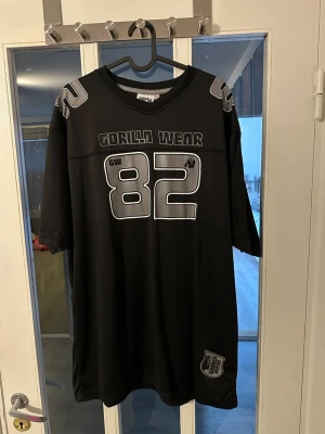 Svart Gorilla Wear t-shirt med tryck - Säljer en svart t-shirt från Gorilla Wear med stort vitt tryck av siffran 82 och logga både fram och bak. T-shirten har korta ärmar, v-ringning och sportig design med detaljer på axlarna. Perfekt för dig som gillar streetwear och sportig stil.