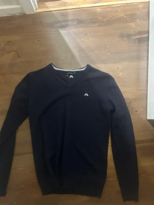   J.Lindeberg tröja - Mörkblå V-ringad tröja från J.Lindeberg med diskret vitt logobrodyr på bröstet. Tröjan har långa ärmar och är tillverkad i mjukt material som känns skönt mot huden. Perfekt för en clean och stilren look. 