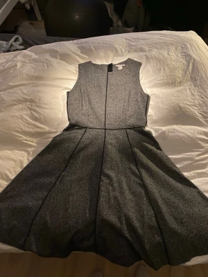 Grå ärmlös klänning med svarta detaljer - Snygg grå ärmlös klänning från HM med svarta sömdetaljer. Klänningen har en lätt utställd kjol och rund halsringning. Materialet känns som en blandning av polyester och bomull, perfekt för dig som gillar stilrena och klassiska plagg. Dragkedja bak på ryggen.