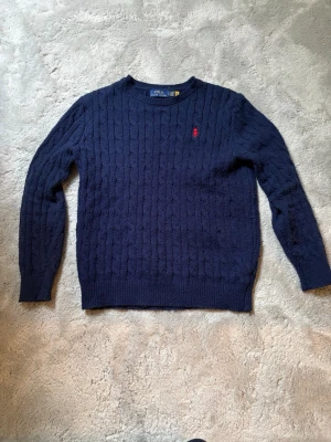 Mörkblå kabelstickad tröja Polo Ralph Lauren - Hör av innan köp! Kabelstickad mörkblå tröja från Polo Ralph Lauren med rund halsringning och röd broderad logga på bröstet. Tröjan har långa ärmar och ribbade muddar vid ärmslut och nederkant. Perfekt för en stilren och klassisk look.