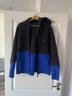 Svart och blå hoodie från BOSS - Snygg hoodie från BOSS i svart och blå färgblock. Dragkedja framtill, justerbar huva med snören och fickor på sidorna. Perfekt för en avslappnad och trendig stil. BOSS-logga broderad på bröstet. Mjuk och bekväm bomullsblandning.