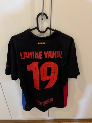 Äkta FC Barcelona Lamine Yamal matchtröja - Säljer en svart FC Barcelona matchtröja från Nike med Lamine Yamal och nummer 19 på ryggen. Tröjan har klubbmärke och Nike-logga framtill, samt röda och blå detaljer på sidorna. Perfekt för fotbollsfans som vill sticka ut på planen eller läktaren.