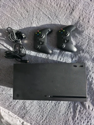 Xbox Series X med två kontroller - Säljer en svart Xbox Series X med två matchande svarta trådlösa handkontroller och tillhörande kablar. Konsolen har en stilren, rektangulär design och är tillverkad i plast. Ingår även flera Xbox One-spel i originalfodral, perfekt för gamingkvällar.