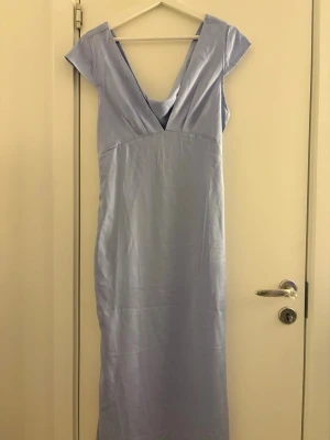 Ljusblå långklänning med v-ringning - Elegant ljusblå långklänning med djup v-ringning både fram och bak. Klänningen har korta ärmar och en slät, figurnära siluett. Perfekt för speciella tillfällen när du vill sticka ut med en stilren look.