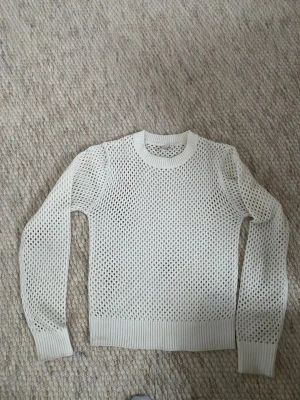 Vit stickad meshtröja - Säljer en vit stickad tröja med meshstruktur och rund hals. Tröjan har långa ärmar och breda ribbade muddar vid ärmslut och nederkant. Perfekt för lager-på-lager och en cool, avslappnad stil.