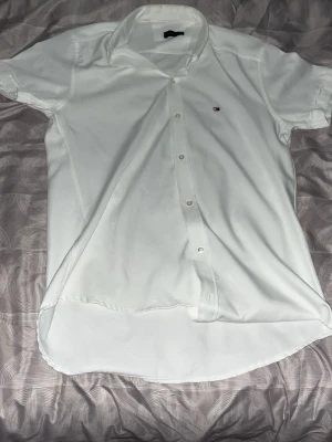 Vit kortärmad skjorta Tommy Hilfiger - Vit kortärmad skjorta från Tommy Hilfiger med klassisk krage och knäppning framtill. Liten broderad logga på bröstet. Skjortan är i ett lätt och svalt material, perfekt för sommaren. Enkel och stilren design som passar till många tillfällen.