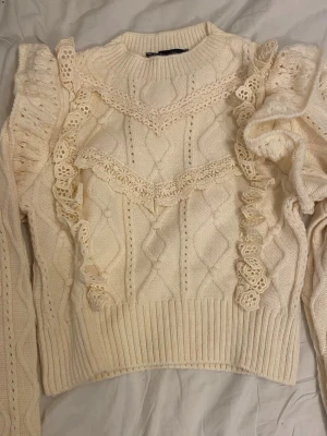 Gullig stickad tröja med volangdetaljer från Mango - Supersöt vit stickad tröja med volangprydda axlar och virkade spetsdetaljer framtill. Tröjan har ett klassiskt kabelstickat mönster, rund halsringning och ribbade muddar. Perfekt för dig som gillar romantisk och trendig stil. Katt finns i hemmet!