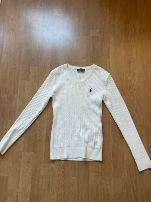 Vit kabelstickad tröja Polo Ralph Lauren - Kabelstickad vit tröja från Polo Ralph Lauren (inte äkta) med V-ringning och broderad marinblå logga på bröstet. Långärmad och ribbstickade muddar. Klassisk och tidlös design i mjukt material. Har en liten fläck på armen men som troligtvis går bort i tvätten. Strl M men skulle definitivt säga att den är mer xs-s
