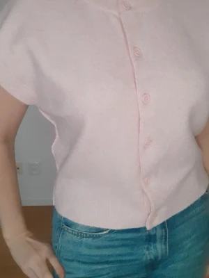 Ljusrosa stickad kortärmad kofta Nelly - Söt ljusrosa stickad kofta med korta ärmar och knappar framtill. Modellen är rak och  perfekt att bära över ett linne eller t-shirt eller som den är. Materiallappen saknas tyvärr, men tyget upplevs som väldigt mjukt, skönt och behagligt mot huden. 