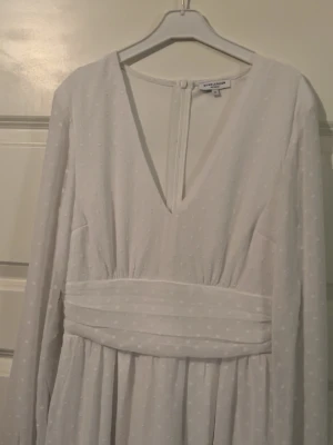 Vit prickig klänning med rosett  - Vit klänning med diskret prickigt mönster, v-ringning och långärmade ärmar. Klänningen har en midjedetalj med en rosett baktill. Tunt, lätt material som ger ett luftigt intryck. Använd bara 2 gånger.