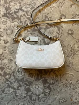 Vit axelväska från Coach - Snygg vit axelväska från Coach med klassiskt monogrammönster och guldfärgade detaljer. Väskan har en smal justerbar axelrem och Coach-logga framtill. Tillverkad i canvas med läderdetaljer och har en tag med Coach-tryck.