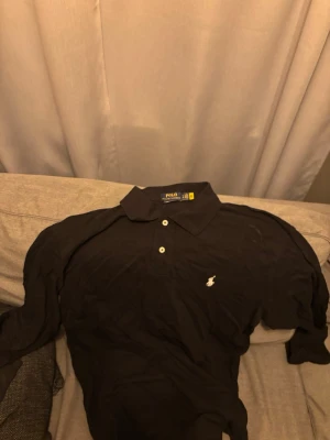 Svart långärmad piké från Polo Ralph Lauren - Klassisk svart långärmad pikétröja från Polo Ralph Lauren med vit broderad logga på bröstet och knappar vid kragen. Skön och tidlös modell i bomull, perfekt till jeans eller chinos.