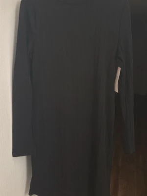 Svart ribbad klänning från Nelly - En svart ribbad klänning från Nelly med lång ärm och rund halsringning. Klänningen är enkel och stilren, perfekt för dig som gillar minimalistisk stil. Materialet är stretchigt och bekvämt.