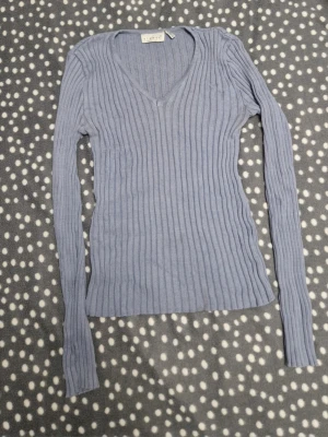 Ljusblå ribbstickad tröja från H&M - Supersnygg ljusblå stickad tröja med ribbad struktur och djup v-ringning. Tröjan har långa ärmar och sitter figurnära, perfekt för lager-på-lager. Materialet känns mjukt och stretchigt.