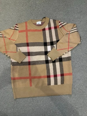 Beige stickad tröja med rutigt mönster - Stickad tröja i beige med klassiskt rutigt mönster i svart, vitt och rött från Burberry. Tröjan har rund halsringning, långa ärmar och ribbade muddar vid ärmslut och nederkant. Perfekt för dig som gillar tidlös och trendig stil.