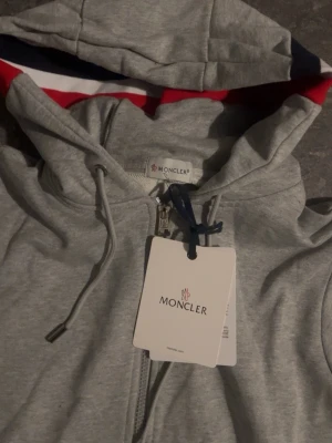 Grå Moncler hoodie med dragkedja - Snygg grå hoodie från Moncler med dragkedja framtill och huva med röd, vit och marinblå insida. Ärmsluten har matchande ränder i samma färger. Justerbar huva med snören och klassisk Moncler-logga på ärmen. Perfekt för en chill och stilren look.